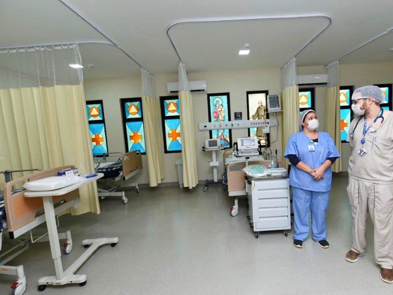 Foto dos novos leitos de terapia intensiva no Hospital Guarapiranga. Do lado esquerdo aparece parte de dois leitos, separados por divisórias de cortina bege. Ao fundo aparece um outro leito na totalidade, com monitores e equipamentos. Mais à frente, do lado direito, dois enfermeiros conversam. Não há pacientes nos leitos